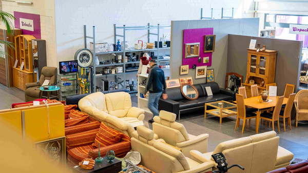 Ein Auktionshaus oder Möbelgeschäft mit verschiedenen Möbeln und Dekorationsgegenständen. Zu sehen sind Sofas, ein Esstisch mit Stühlen und Regale im Hintergrund. Menschen bewegen sich zwischen den Möbeln. (Text KI generiert)