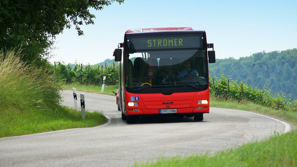 Ein roter Stadtbus fährt auf einer kurvenreichen Landstraße. Die Anzeige auf dem Bus weist die Strecke "STROMER" aus. Im Hintergrund sind grüne Felder und Bäume sichtbar, die eine ländliche Umgebung darstellen.