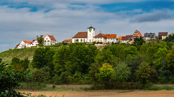 Ein malerisches Dorf liegt auf einem Hügel, umgeben von Wäldern und Weinbergen. Die Hauptattraktion ist die Kirche mit einem Uhrturm. Die Häuser sind in warmen Farbtönen gehalten, was eine einladende Atmosphäre schafft. Der Himmel zeigt einen leicht bewölkten Tag.