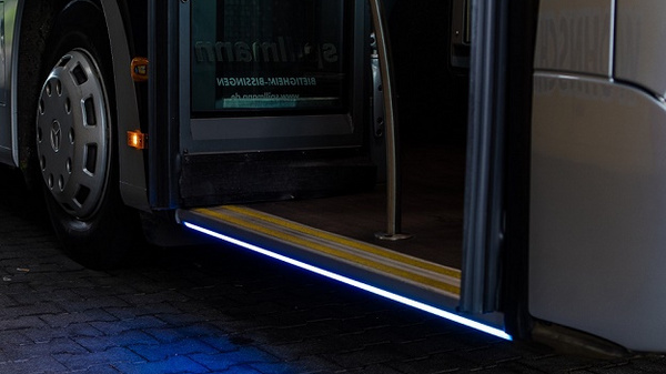 A close-up view of a bus entrance with its door open, showcasing a lit step with glowing blue and yellow highlights. The bus is parked on a cobblestone surface, emphasizing the accessibility of the entrance.