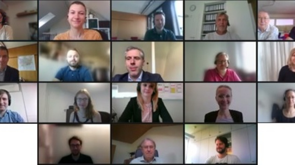 Une grille de visages de participants lors d'une réunion en ligne. Différents individus sourient et regardent vers l'écran, chacun dans son propre espace. L'image montre une diversité de personnes engagées dans une conversation virtuelle.