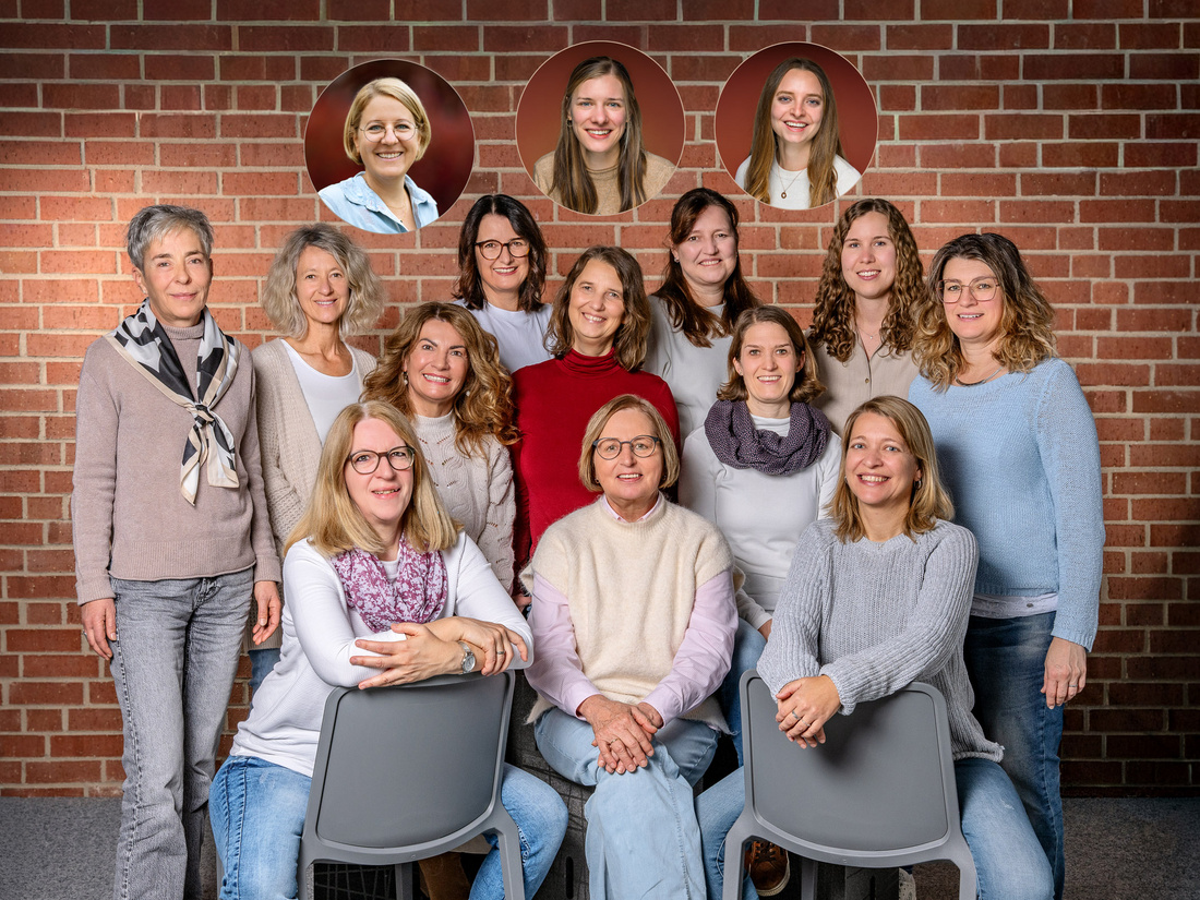 Eine Gruppe von 15 Frauen posiert vor einer Backsteinwand. Einige Frauen sitzen auf Stühlen, andere stehen dahinter. In Kreisen über der Gruppe sind Porträts von vier weiteren Frauen abgebildet. Alle Frauen lächeln freundlich in die Kamera. (Text KI generiert)