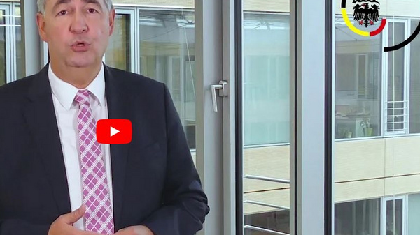 A man in a suit stands in an office space with large windows, speaking directly to the camera. He gestures with his hands as he presents information, while a red YouTube play button appears in the center of the image, indicating a video format.