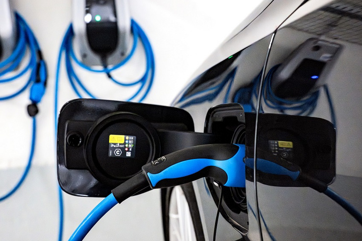 Ein Elektroauto wird an einer Ladestation aufgeladen. Die Ladebuchse des Fahrzeugs ist geöffnet, und ein Kabel mit blauem Griff ist eingesteckt. Im Hintergrund sind Ladestationen zu sehen, die ebenfalls mit Kabeln verbunden sind.