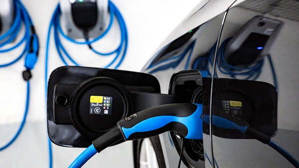Ein Elektroauto wird an einer Ladestation aufgeladen. Die Ladebuchse des Fahrzeugs ist geöffnet, und ein Kabel mit blauem Griff ist eingesteckt. Im Hintergrund sind Ladestationen zu sehen, die ebenfalls mit Kabeln verbunden sind.