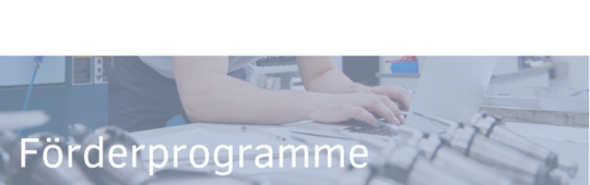 Ein Mitarbeiter arbeitet am Computer und ist mit technischen Geräten umgeben. Der Text „Förderprogramme“ ist deutlich sichtbar. Die Bildkomposition vermittelt ein industrielles Arbeitsumfeld, das sich auf Förderprogramme konzentriert.