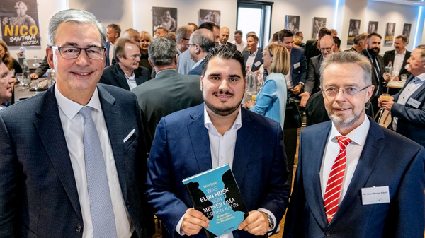 Trois hommes posent au premier plan d'un événement, entourés de personnes en arrière-plan. L'un des hommes tient un livre intitulé "Elon Musk : Mein Leben". Les autres semblent engagés dans une conversation, dans une ambiance de réunion professionnelle.
