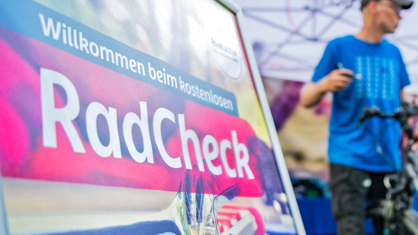 Ein Schild mit der Aufschrift "Willkommen beim kostenlosen RadCheck" steht im Vordergrund. Im Hintergrund sind zwei Männer zu sehen, die Fahrräder überprüfen. Einer der Männer spricht, während der andere an einem Fahrrad arbeitet. Es herrscht eine freundliche, einladende Atmosphäre.