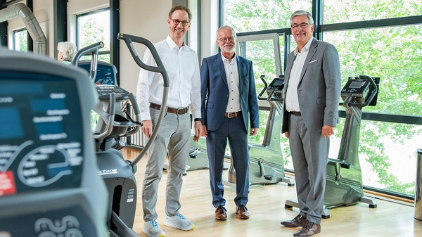 Drei Männer stehen in einem Fitnessstudio, umgeben von Fitnessgeräten. Sie lächeln und wirken freundlich. Die Umgebung ist hell und modern, mit großen Fenstern, die Tageslicht hereinlassen.