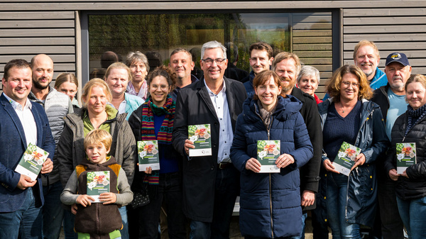 Vor einem Gebäude mit dem Schriftzug "Hofladen" steht eine Gruppe von etwa 20 Personen. Sie halten Broschüren in der Hand und lächeln. Die Szene vermittelt eine freundliche Atmosphäre und zeigt eine Gemeinschaft, die sich für lokale Produkte engagiert.