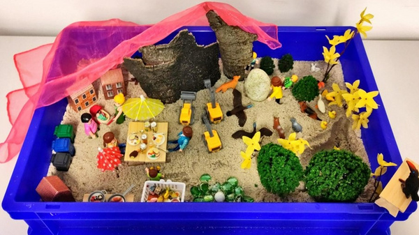 Eine kreative Spielszene in einer blauen Box mit Sand. Figuren sitzen an einem Tisch unter einem Sonnenschirm, während andere spielen oder arbeiten. Es gibt Bäume, Blumen, und Spielzeugfahrzeuge. Ein pinker Stoff hängt als dekoratives Element über der Szene.