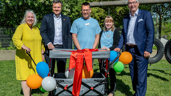 Fünf Personen stehen fröhlich auf einer Wiese. Sie lächeln und umarmen einen Kinderwagen, der mit bunten Ballons geschmückt ist. Die Szene vermittelt eine feierliche Atmosphären, möglicherweise anlässlich einer Veranstaltung oder einer Spendenaktion für Kinder.