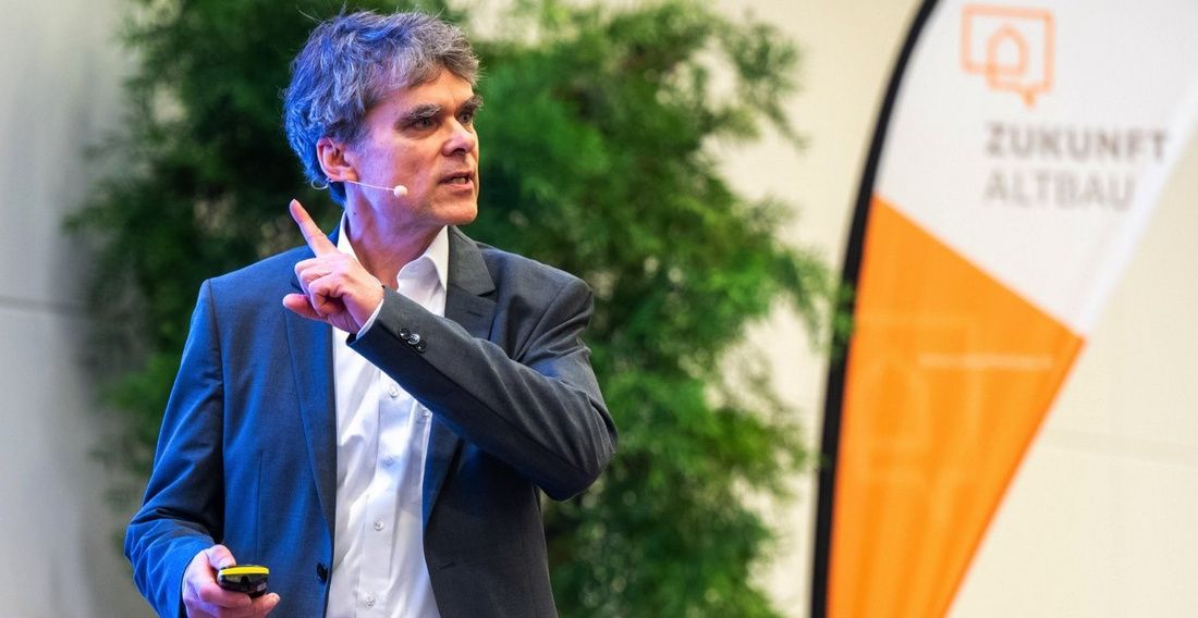 Ein Referent steht auf einer Bühne und spricht, während er einen Zeigestock in der Hand hält. Im Hintergrund ist ein Banner mit dem Text "Zukunft Altbau" zu sehen. Die Szene vermittelt eine Präsentation oder Vorlesung zu einem Thema im Bereich Altbausanierung.