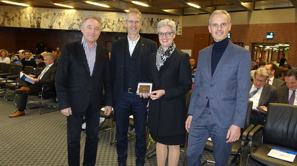 Four individuals are standing together in a conference room, holding a small plaque. They appear to be engaged in a formal event, with an audience visible in the background. The setting features chairs and a detailed mural on the wall above.
