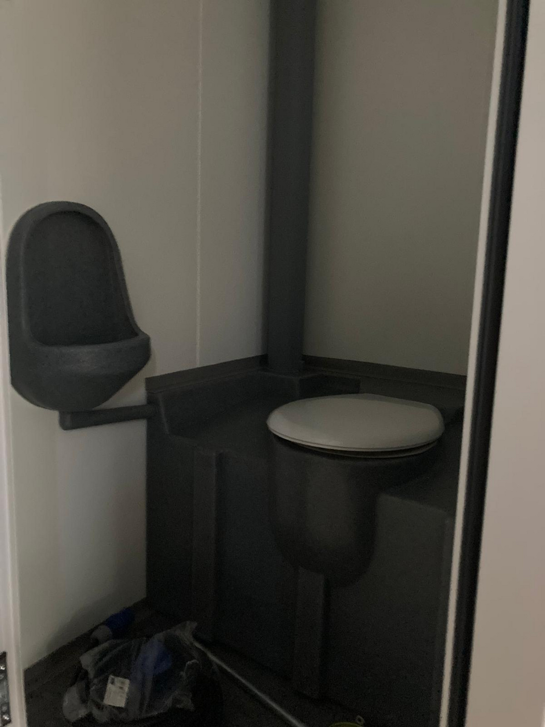 Das Bild zeigt eine einfache Toiletteneinheit mit einem Wandurinal auf der linken Seite und einer Toilette auf der rechten Seite. Der Raum hat grau-weiße Wände und einen minimalistischen, funktionalen Stil.