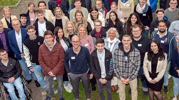 Un gruppo di circa quaranta persone è riunito in un'area verde all'aperto. Ci sono uomini e donne di diverse età, alcuni in abiti formali e altri casual. Alcune persone sono sorridenti e sembra che stiano partecipando a un evento di gruppo o un incontro.
