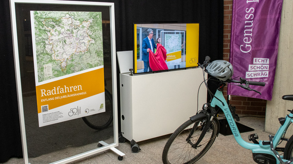 Eine Ausstellung über Radfahren mit einem großen Infobreich, der eine Karte zeigt. Neben dem Infobereich stehen Fahrräder und ein Präsentationsbildschirm. In der Nähe befinden sich Informationsbroschüren zum Thema Radfahren. Im Hintergrund sind dekorative Displays zu sehen.