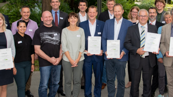 A diverse group of individuals stands together outdoors, smiling and holding certificates. They represent a variety of professional backgrounds and are positioned in two rows against a backdrop of greenery. The atmosphere is celebratory, indicating a recognition or achievement event.