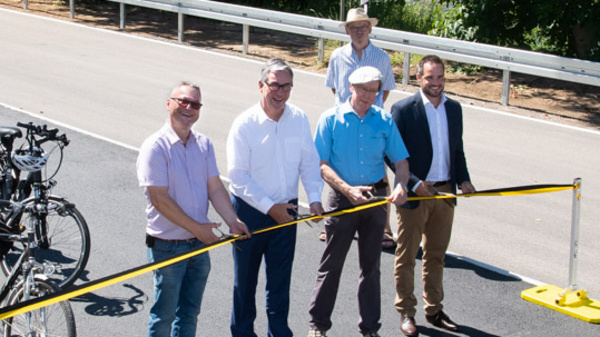 A group of eight people, some with bicycles, gather on a newly paved road. They stand behind a yellow ribbon stretched across the road, symbolizing the opening of a cycling path. Trees and a roadside sign are visible in the background.