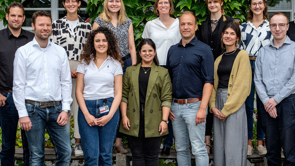 Eine Gruppe von 15 Personen steht vor einer mit Efeu bewachsenen Wand. Sie posieren gemeinsam, einige lächeln. Die Gruppe besteht aus Männern und Frauen unterschiedlichen Alters, die leger gekleidet sind.