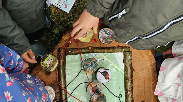 Eine Gruppe von Kindern betrachtet ein interaktives Spiel auf einem Holzbrett. Auf dem Brett sind runde Plättchen mit verschiedenen Bildern angeordnet, die durch Schnüre miteinander verbunden sind. Die Kinder scheinen aktiv am Spielen und Entdecken zu sein.
