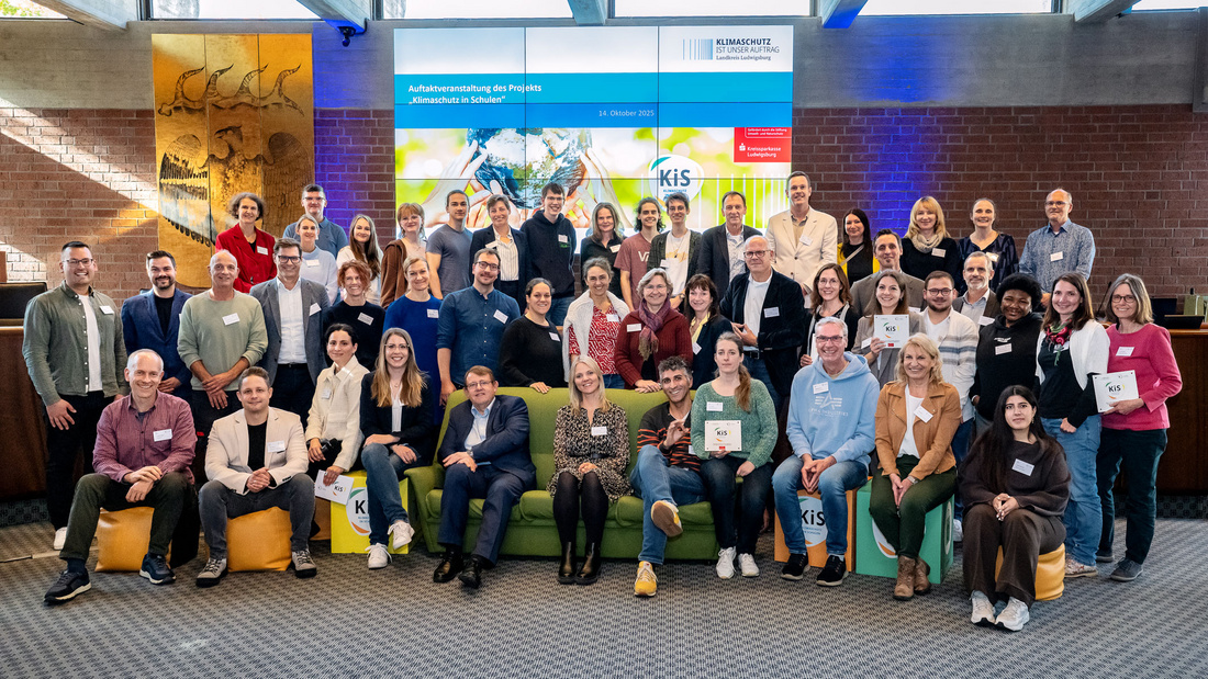 Eine große Gruppe von Menschen steht und sitzt zusammen in einem Raum. Sie halten Urkunden oder Zertifikate in den Händen und lächeln freundlich. Im Hintergrund ist ein Bildschirm mit Informationen zu sehen. Die Veranstaltung scheint eine Feier oder Auszeichnung zu sein.