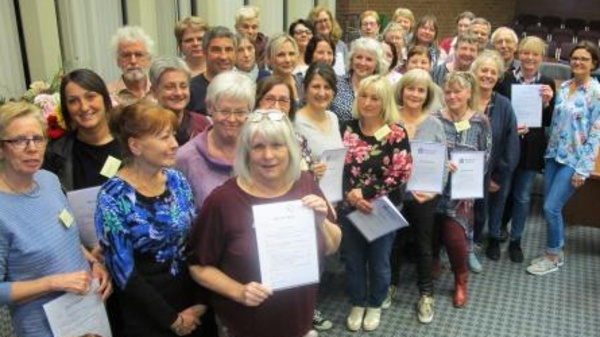 A large group of people stands together, smiling and holding certificates. They are gathered indoors, with a mix of men and women of various ages, indicating a celebration or recognition event. A few individuals are positioned in the front, showcasing their certificates prominently.