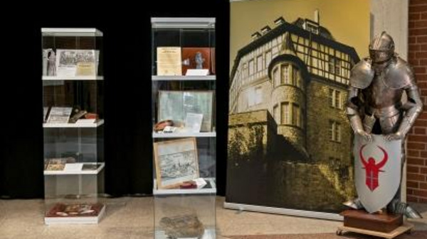 The image features a display area showcasing historical artifacts. On the left, glass cases hold various items and documents. To the right, a large banner depicts a building, while a suit of armor stands next to it, holding a shield with a red emblem.