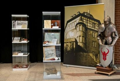Eine Ausstellung mit Vitrinen, die historische Dokumente und Objekte zeigen, steht neben einem Banner mit einer historischen Gebäudeansicht. Rechts steht eine Rüstung mit einem Schild, der ein rotes Symbol zeigt. Der Hintergrund ist dunkel, um die Exponate hervorzuheben.