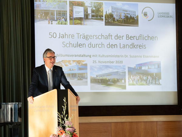 25.11.2020 - 50 Jahre Trägerschaft der Beruflichen Schulen,Veranstaltung an der Carl-Schaefer-Schule Eine Person steht an einem Holzpult und spricht. Im Hintergrund befindet sich eine Leinwand mit der Aufschrift „50 Jahre Trägerschaft der Beruflichen Schulen durch den Landkreis“ sowie weiteren Informationen zur Veranstaltung. Das Datum ist der 25. November 2020.