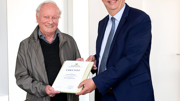 Zwei Männer stehen zusammen in einem hellen Raum. Der Mann links, älter, trägt einen grauen Pullover und hält eine Urkunde. Der Mann rechts, in Anzug und Krawatte, überreicht ihm die Urkunde lächelnd. Beide zeigen Freude über den Moment des Anerkennens.