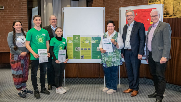 Un groupe de six personnes se tient devant un panneau d'affichage. Elles tiennent des certificats et sourient. Le panneau contient des informations et des graphiques, probablement liées à un projet ou une présentation. L'occasion semble festive et de célébration.