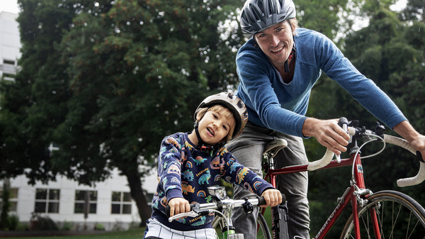Ein Mann und ein Junge stehen neben ihren Fahrrädern in einem grünen Park. Beide tragen Helme und lächeln. Der Junge sitzt auf seinem kleinen Fahrrad, während der Mann ihn freundlich ansieht. Im Hintergrund sind Bäume und ein Gebäude sichtbar.