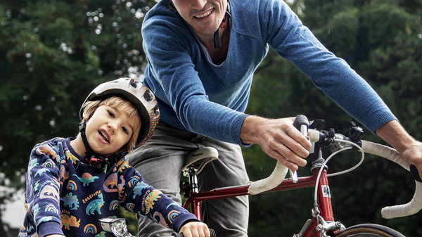 Un homme et un enfant souriant portent des casques de vélo. L'homme aide l'enfant à se préparer à faire du vélo. Ils sont dans un parc, entourés d'arbres. L'atmosphère est joyeuse et conviviale.