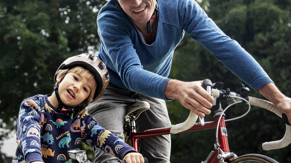 Ein Mann und ein Junge stehen neben ihren Fahrrädern in einem grünen Park. Beide tragen Helme und lächeln. Der Junge sitzt auf seinem kleinen Fahrrad, während der Mann ihn freundlich ansieht. Im Hintergrund sind Bäume und ein Gebäude sichtbar.