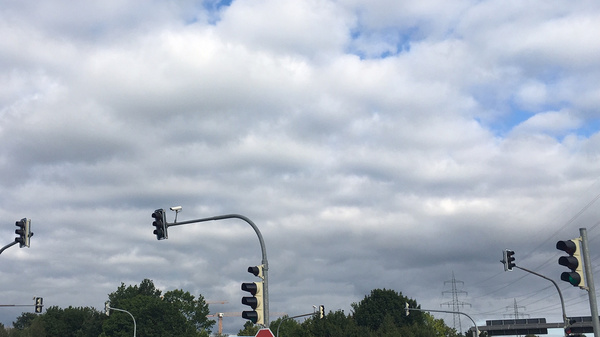 A busy intersection is depicted with several vehicles stopped at traffic lights. The sky is overcast with clouds. Traffic signals include green and red lights, indicating movement and stopping for cars. A stop sign is also visible, guiding drivers at the intersection.