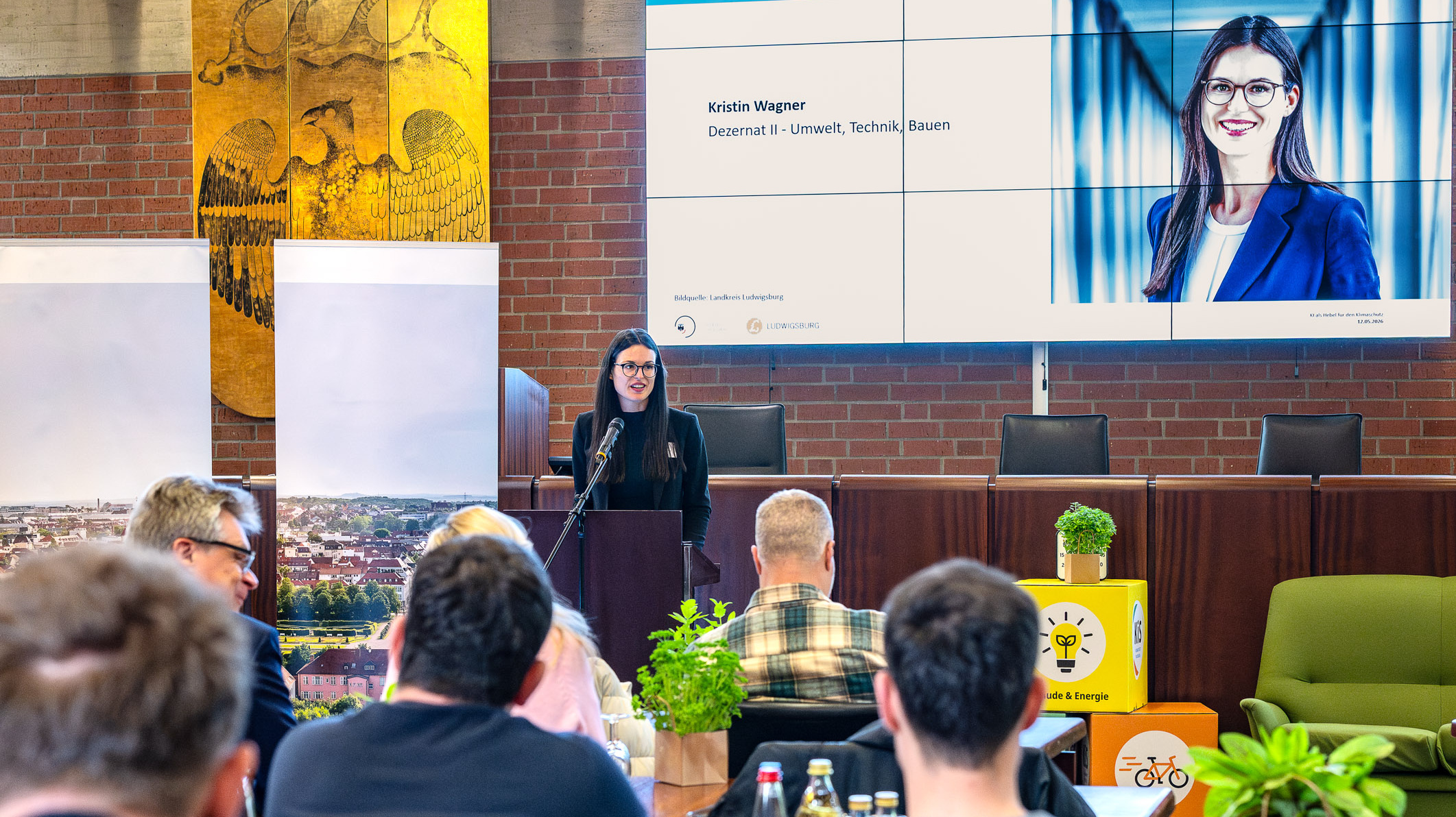 Eine Frau steht hinter einem Podium und spricht zu einem Publikum. Im Hintergrund ist eine große Leinwand mit ihrem Namen und Beruf zu sehen. Der Raum ist mit Pflanzen und Getränken dekoriert, und ein Stadtbild ist sichtbar.