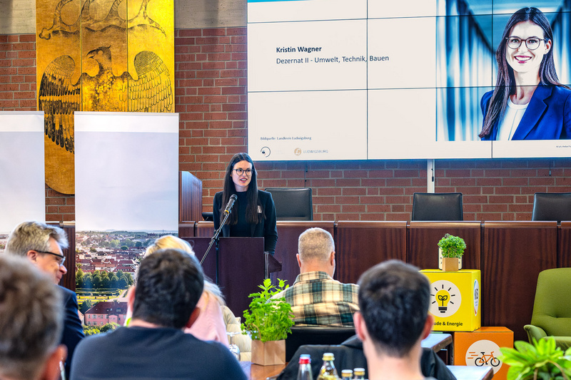 Eine Frau steht hinter einem Podium und spricht zu einem Publikum. Im Hintergrund ist eine große Leinwand mit ihrem Namen und Beruf zu sehen. Der Raum ist mit Pflanzen und Getränken dekoriert, und ein Stadtbild ist sichtbar.