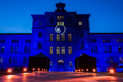 Ein historisches Gebäude ist bei Nacht in blauem Licht beleuchtet. In der Mitte der Fassade ist ein rundes Logo projiziert. Vor dem Gebäude stehen zwei schwarze Zelte und Scheinwerfer sorgen für eine festliche Atmosphäre.