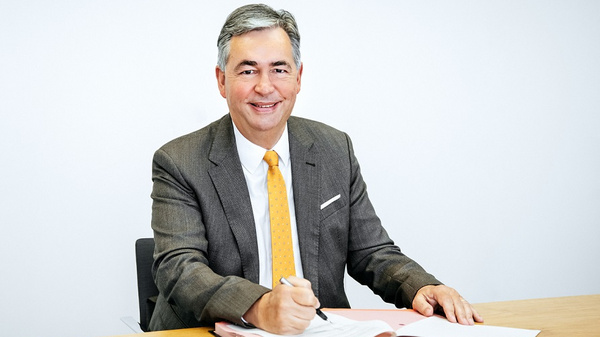 A professional man sits at a wooden table, smiling as he writes in a notebook. He is dressed in a grey suit and orange tie, exuding a confident and approachable demeanor. The background is plain and neutral, emphasizing the individual.