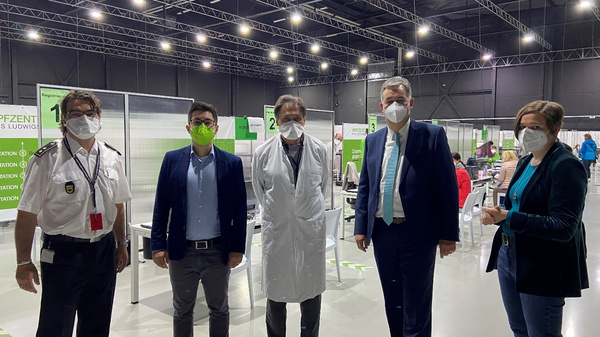 A group of five individuals, including healthcare workers and officials, are standing in a large, well-lit space set up for vaccinations. All are wearing face masks. The backdrop features clear signs indicating a vaccination site, with partitions separating different areas.