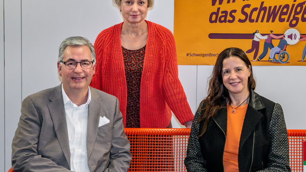Drei Personen sitzen auf einer orangen Bank in einem Raum mit einem Plakat im Hintergrund. Das Plakat trägt den Text „Wir brechen das Schweigen“ und thematisiert den Internationalen Tag gegen Gewalt an Frauen am 25. November.