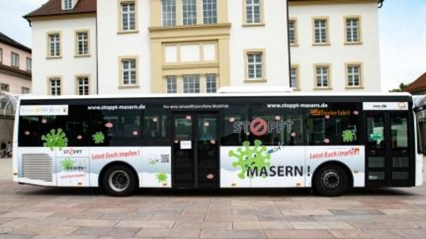 A public bus features a campaign against measles, displaying the message "STOPP MASERN!" along with encouraging text "Lasst Euch impfen!" in bright colors. The bus is parked in front of a building, promoting vaccination awareness.