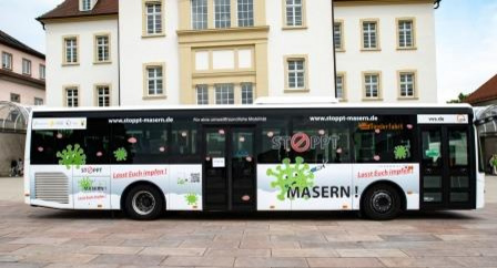 A public bus features a campaign against measles, displaying the message "STOPP MASERN!" along with encouraging text "Lasst Euch impfen!" in bright colors. The bus is parked in front of a building, promoting vaccination awareness.