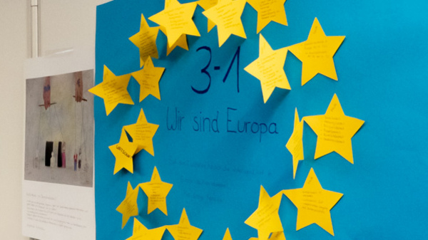 The image shows a display board featuring children's artwork, including colorful drawings. Prominently featured is a blue poster with yellow stars arranged in a circle, displaying the text "3=1 Wir sind Europa," highlighting a theme of unity in Europe.