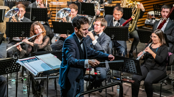Ein Dirigent steht an der Spitze eines Orchesters, das aus mehreren Musikern besteht. Die Musiker spielen verschiedene Instrumente, darunter Bläser und Streicher. Der Dirigent hat seinen Taktstock in der Hand und leitet die Aufführung mit Energie und Leidenschaft.