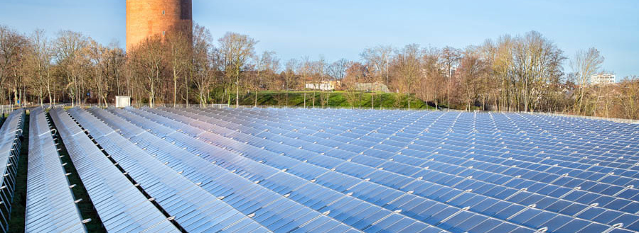 Header Bild: Eine große Fläche mit Solarpanelen erstreckt sich im Vordergrund, während im Hintergrund ein historischer Wasserturm steht. Der Himmel ist blau und klar, und es sind einige Bäume in der Umgebung zu sehen. Die Szenerie vermittelt eine Atmosphäre von nachhaltiger Energieproduktion.