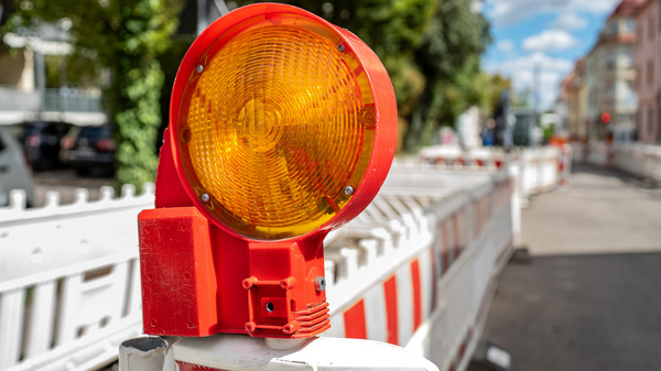 Eine orangefarbene Baustellenlampe steht auf einem weißen Absperrzaun. Im Hintergrund sind Bäume und eine Straße mit Autos zu sehen. Der Himmel ist blau mit einigen Wolken. (Text KI generiert)