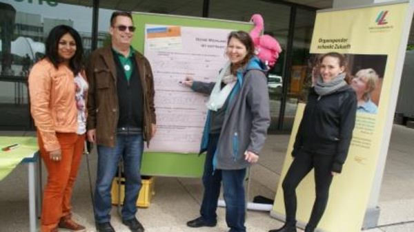 Four people stand near an information board outdoors, engaged in discussion. Two women and two men are present, with one woman pointing at the board. Informational banners are visible in the background, promoting future opportunities. The setting appears to be part of a community or organizational event.