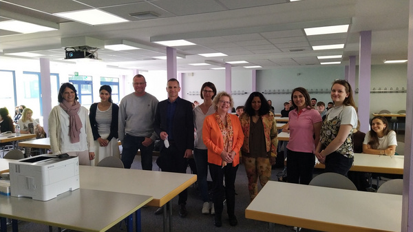 Eine Gruppe von neun Personen steht in einem Klassenzimmer. Einige Personen tragen formelle und andere legere Kleidung. Im Hintergrund sind Tische und Stühle vorhanden. Es scheint ein Treffen oder eine Veranstaltung zu sein.